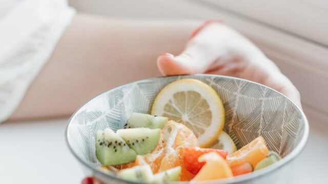 crop woman having bowl of fresh fruit salad at windowsill